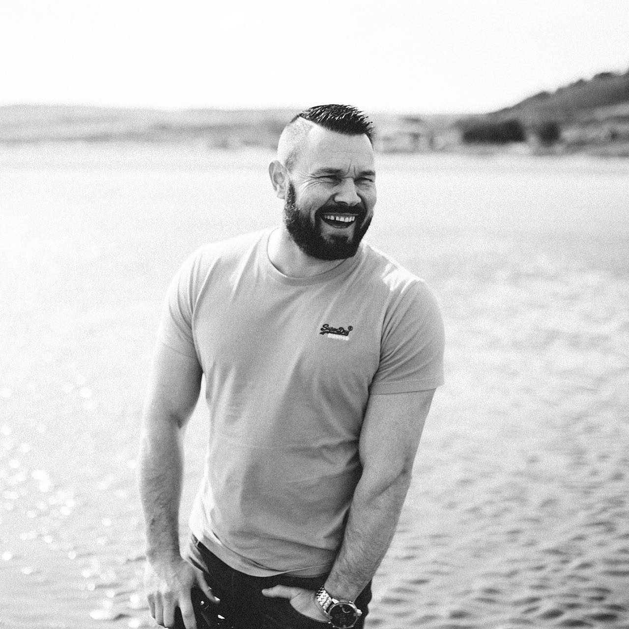 A Black & White portrait of Swansea Wedding Photographer. He stands Laughing at something. Photograph Taken on a south wale beach.